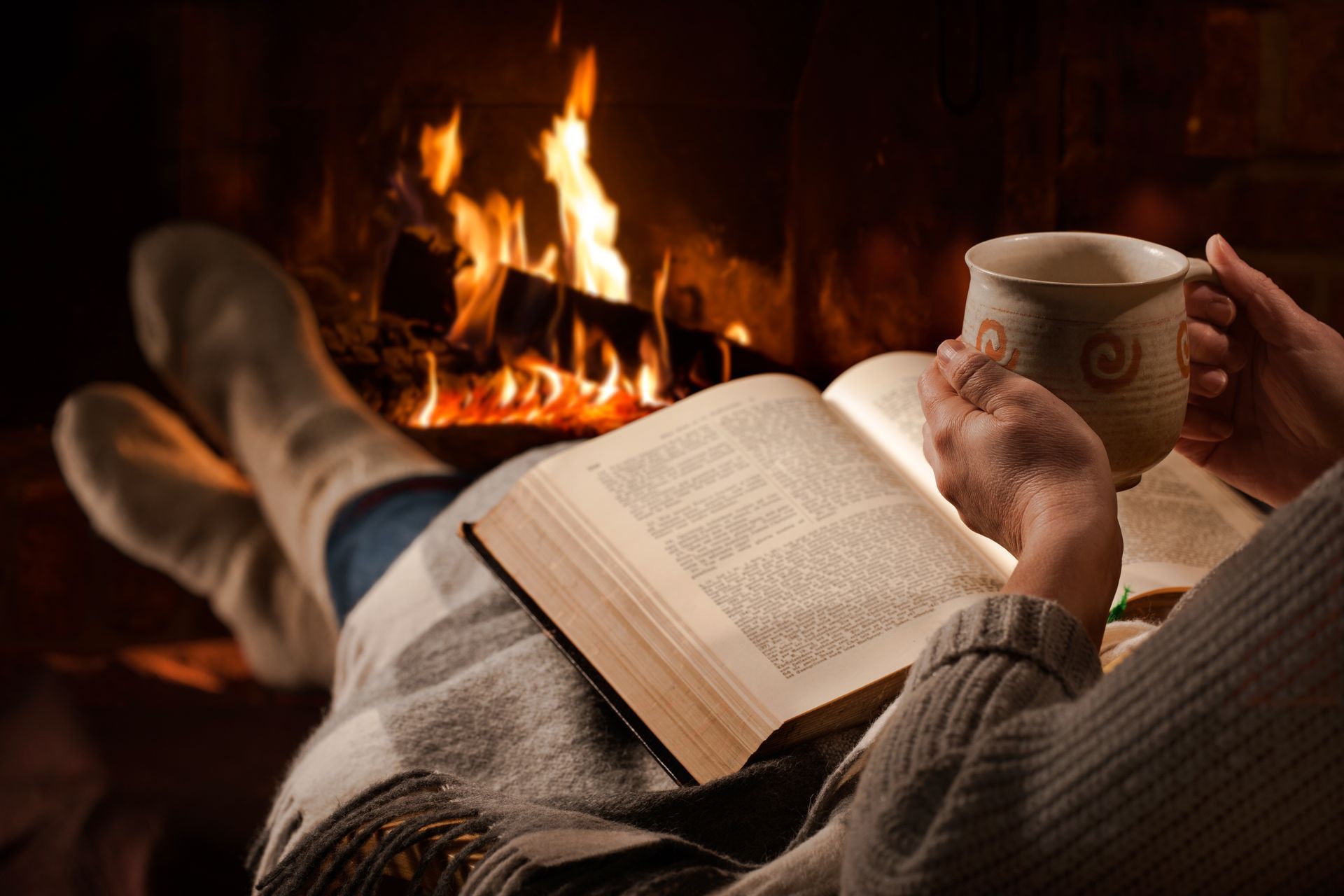 A person is reading a book and drinking coffee in front of a fireplace.