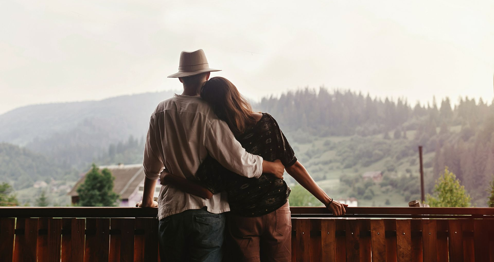 Couple on a balcony, embracing while looking at a mountain range. Man in hat. Cloudy sky.
