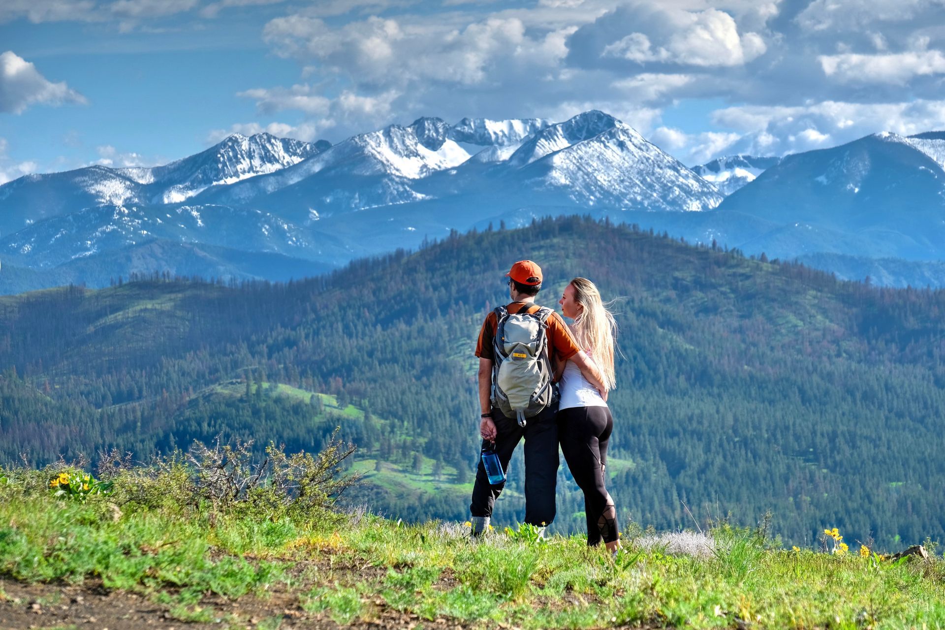 A man and a woman are standing on top of a hill looking at the mountains.