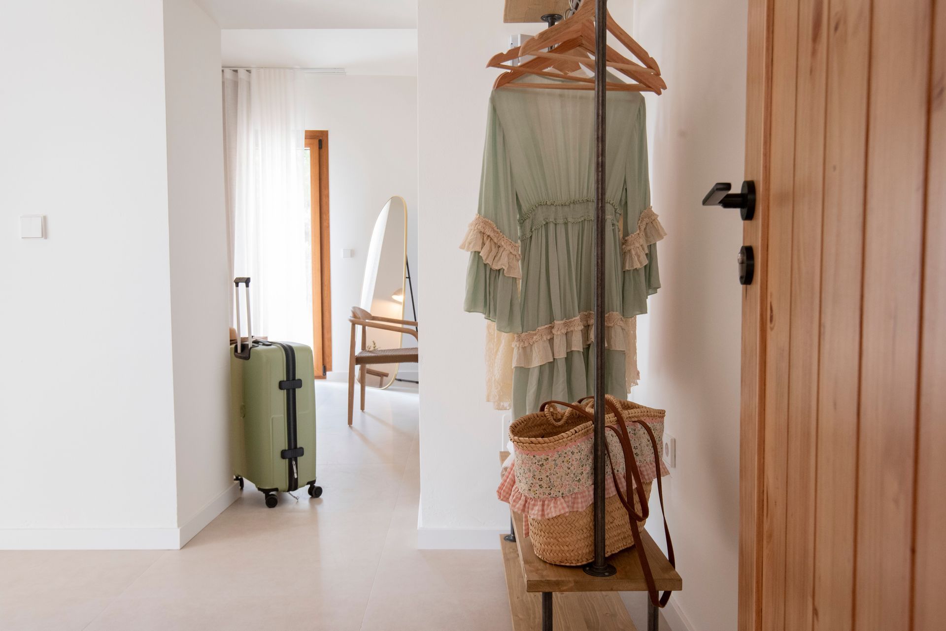 An olive green suitcase, a green dress, and a straw bag hang in a white-walled hallway.