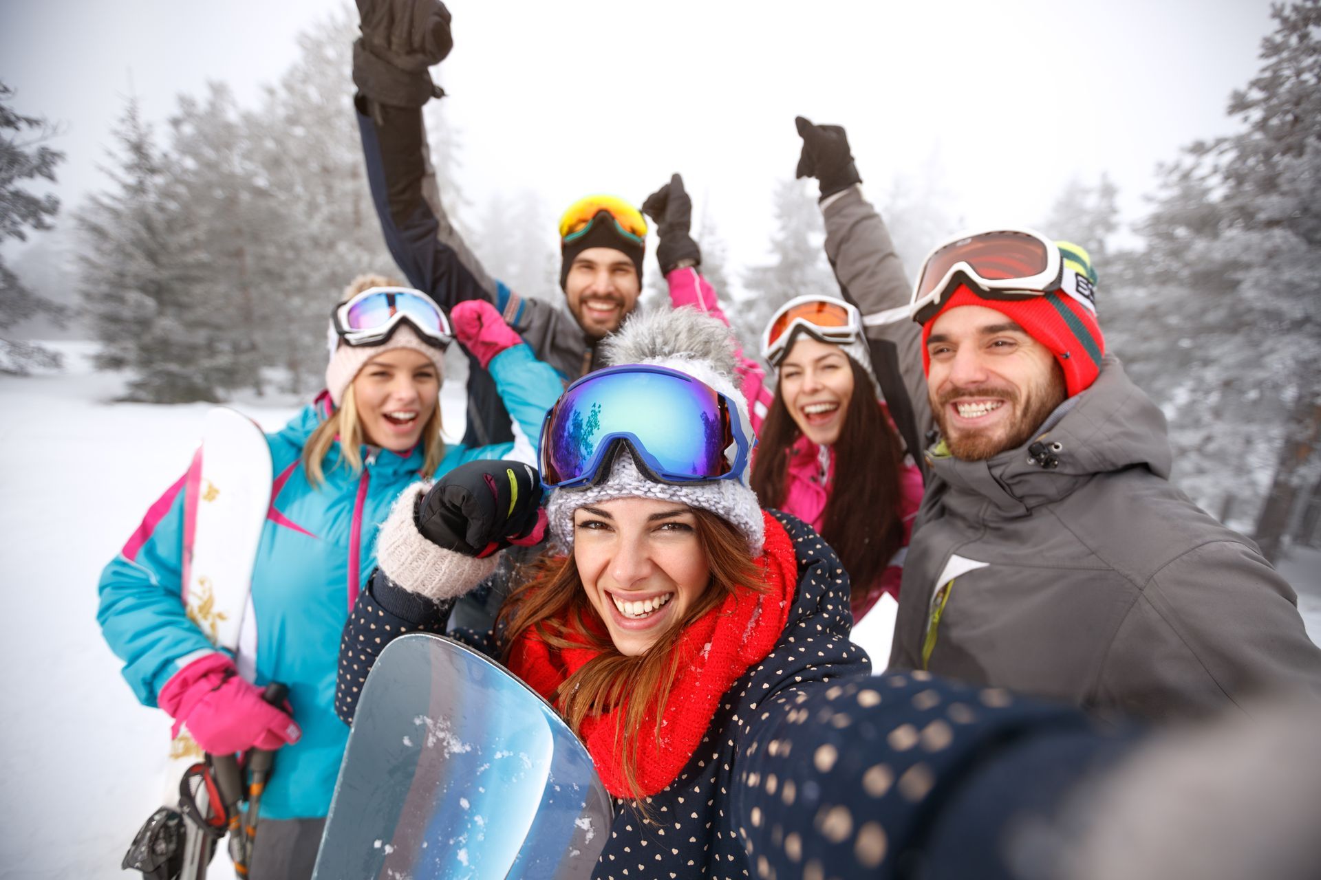 Group of friends in winter gear pose for selfie in snowy landscape, smiling, raising arms.