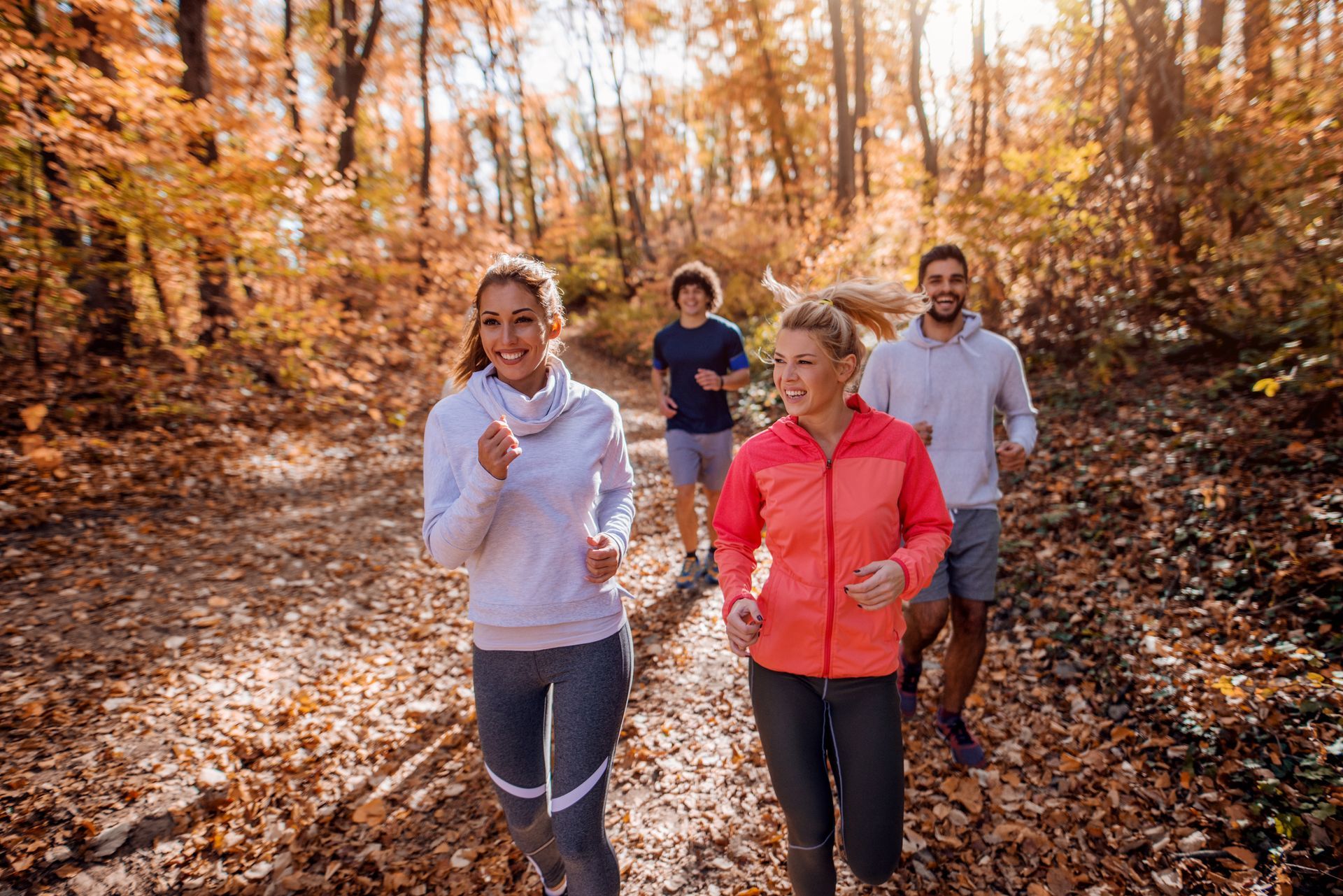 Four people running on a trail in a colorful autumn forest. They are smiling and wearing athletic clothes.