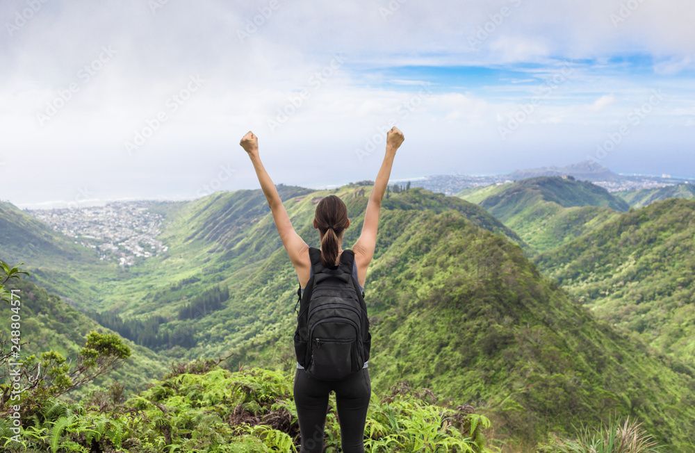 A woman with a backpack is standing on top of a mountain with her arms in the air.