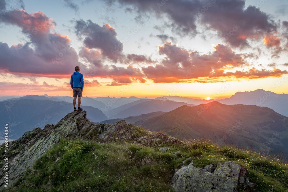 A man is standing on top of a mountain looking at the sunset.