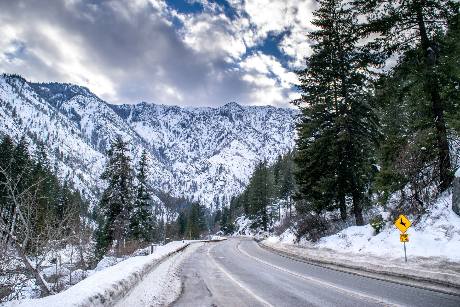 A snowy road with mountains in the background and trees on the side.