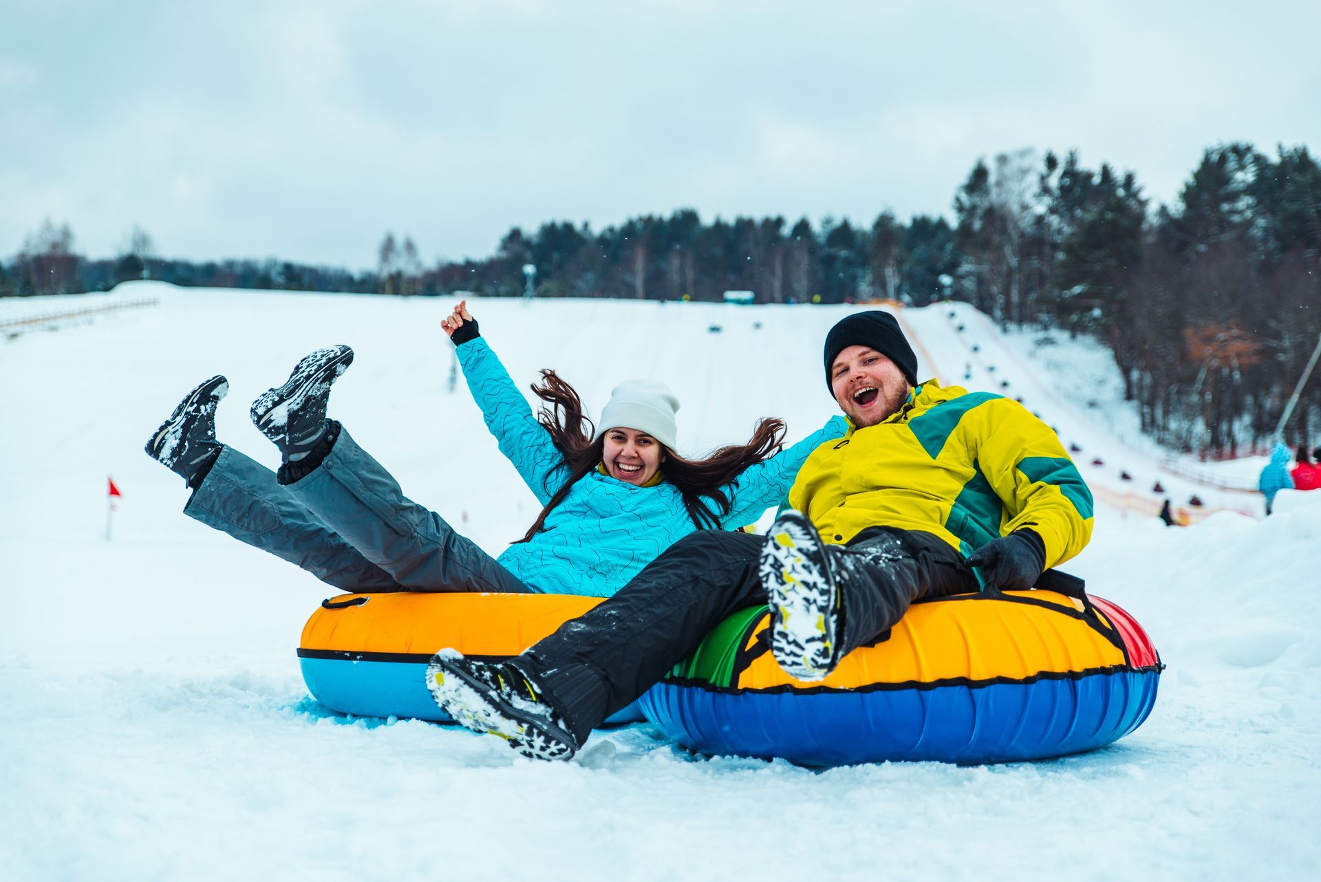 Two people tubing down a snowy hill, smiling, and raising arms in excitement.