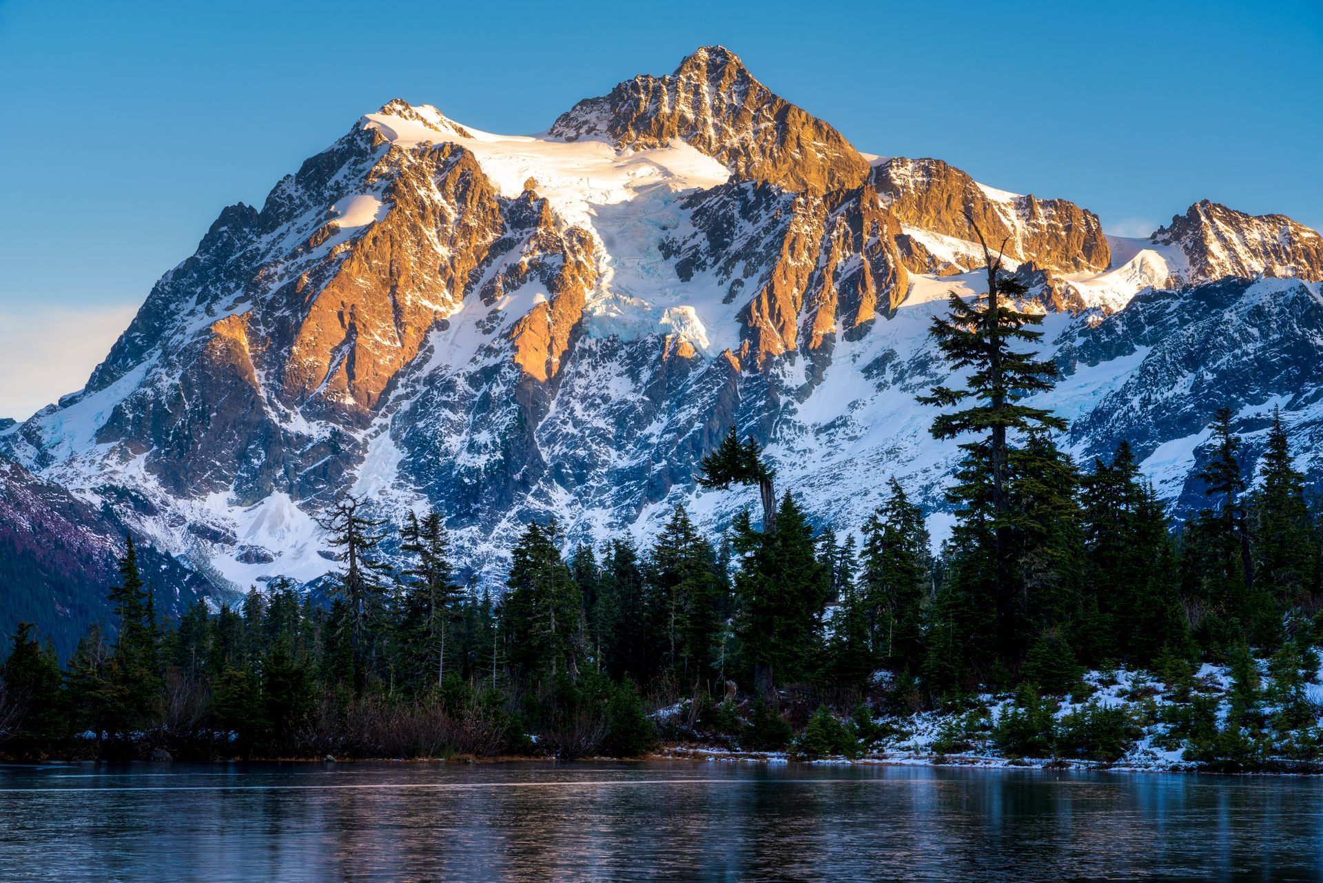 A mountain covered in snow is surrounded by trees and a lake.