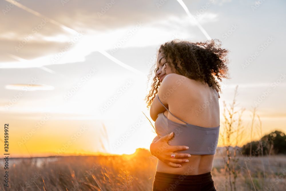 A woman is hugging herself in a field at sunset.