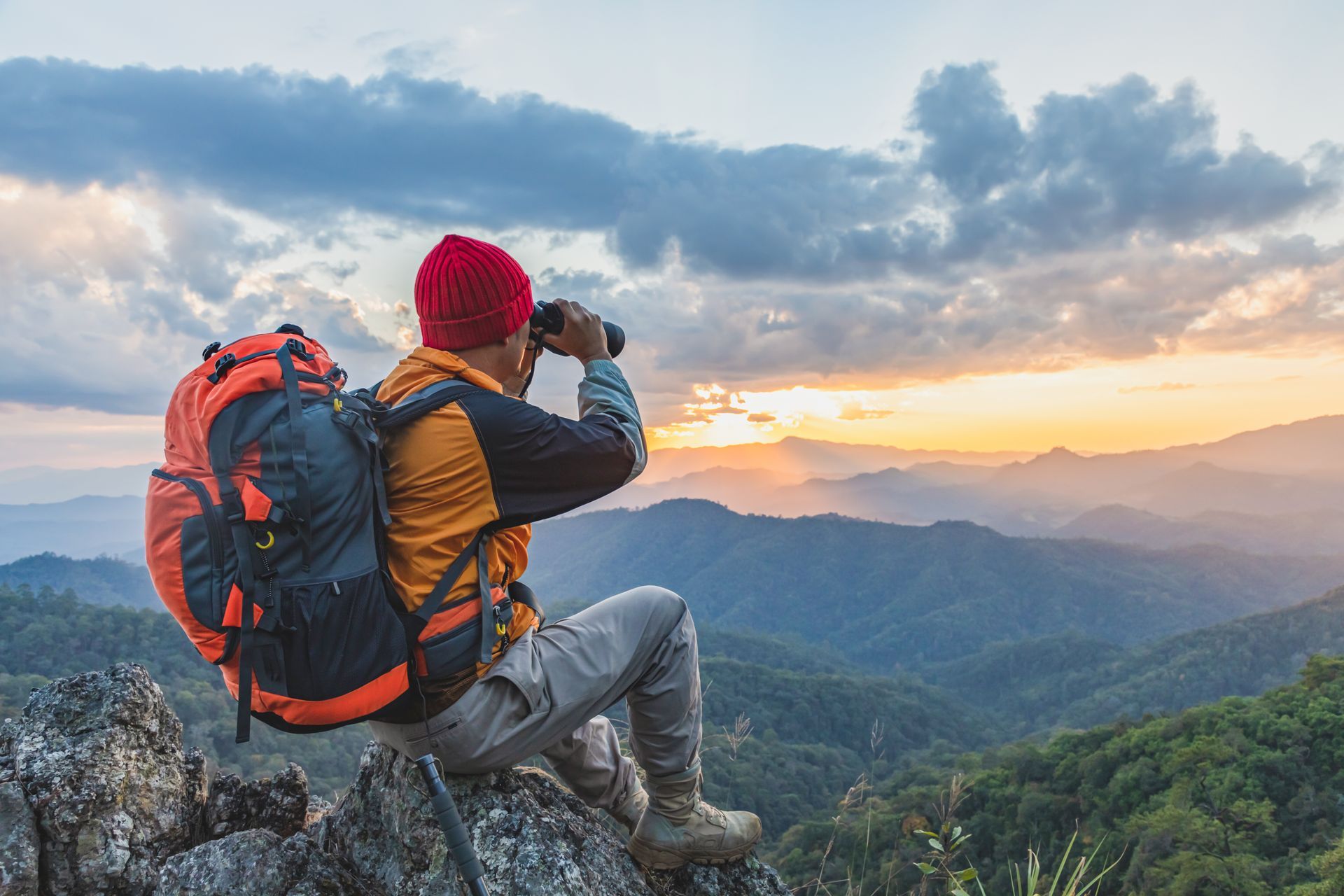 A man with a backpack is sitting on top of a mountain looking through binoculars.