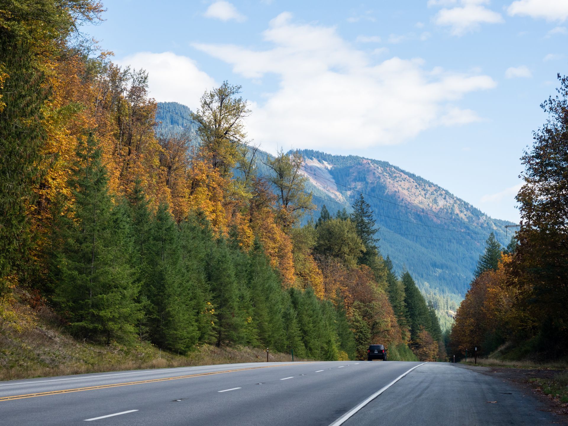 Highway through colorful autumn trees and a mountain under a blue sky.