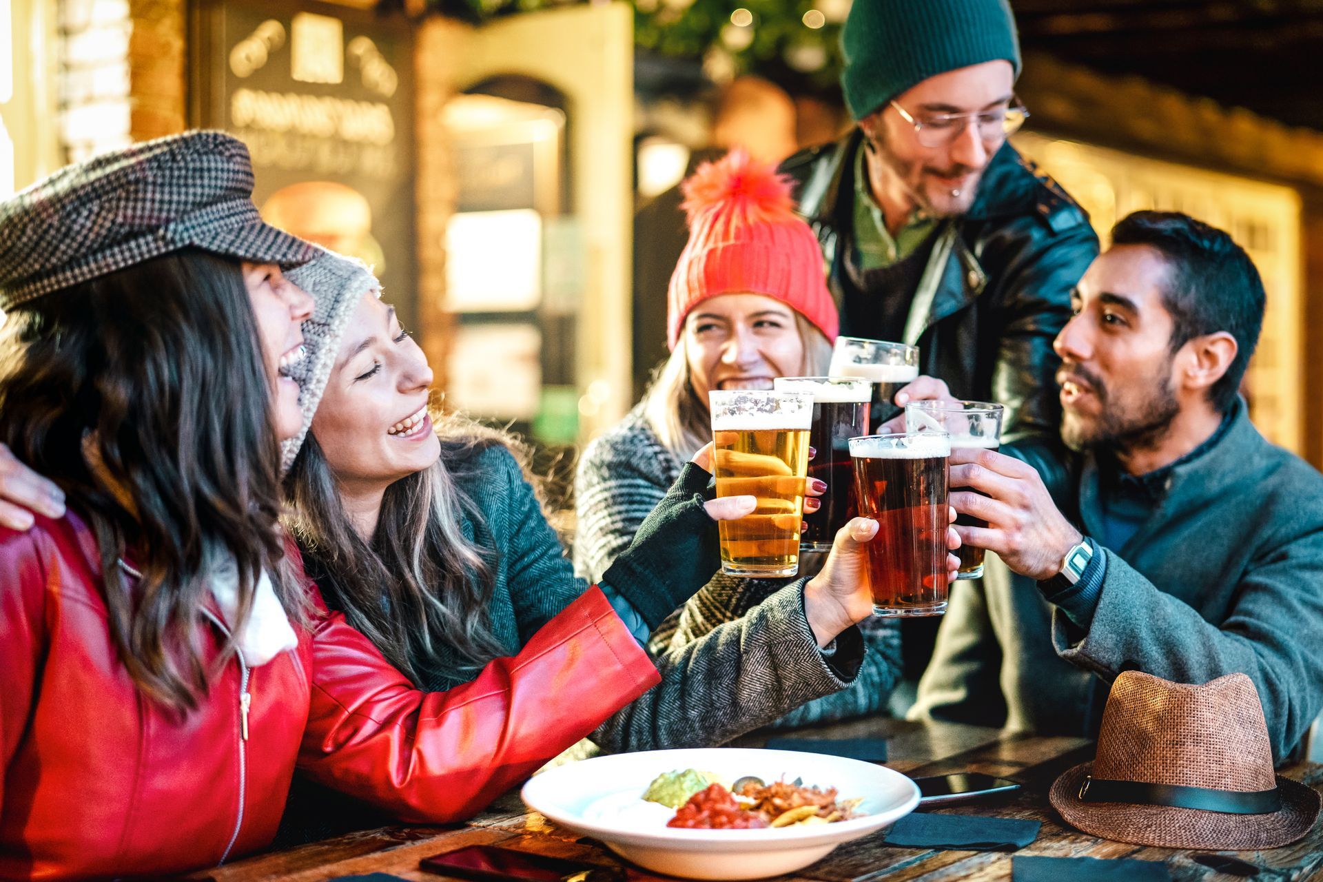 Group of friends clinking beer glasses at an outdoor table, smiling and laughing.