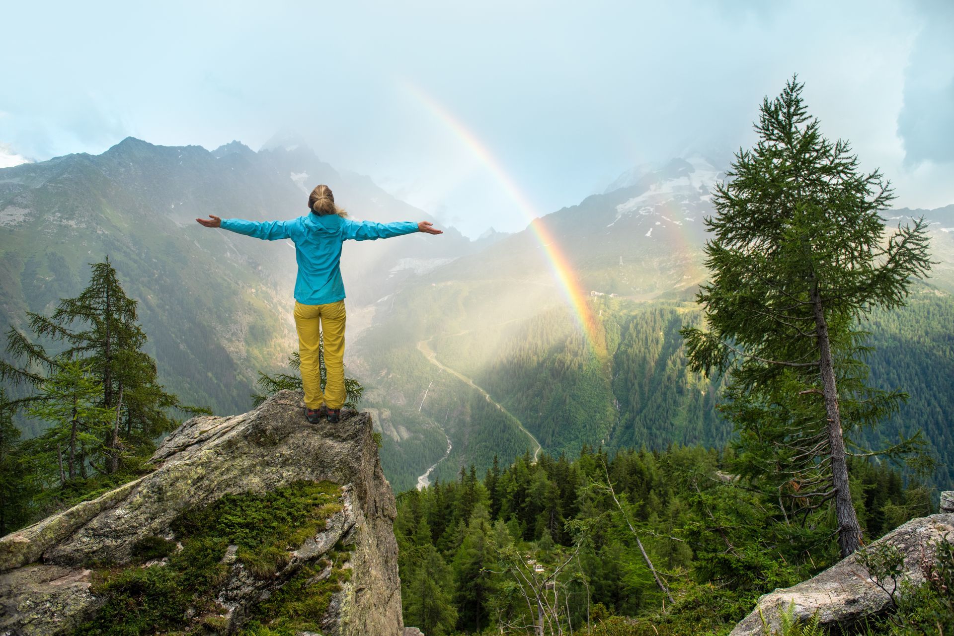 A woman is standing on top of a mountain with her arms outstretched and a rainbow in the background.