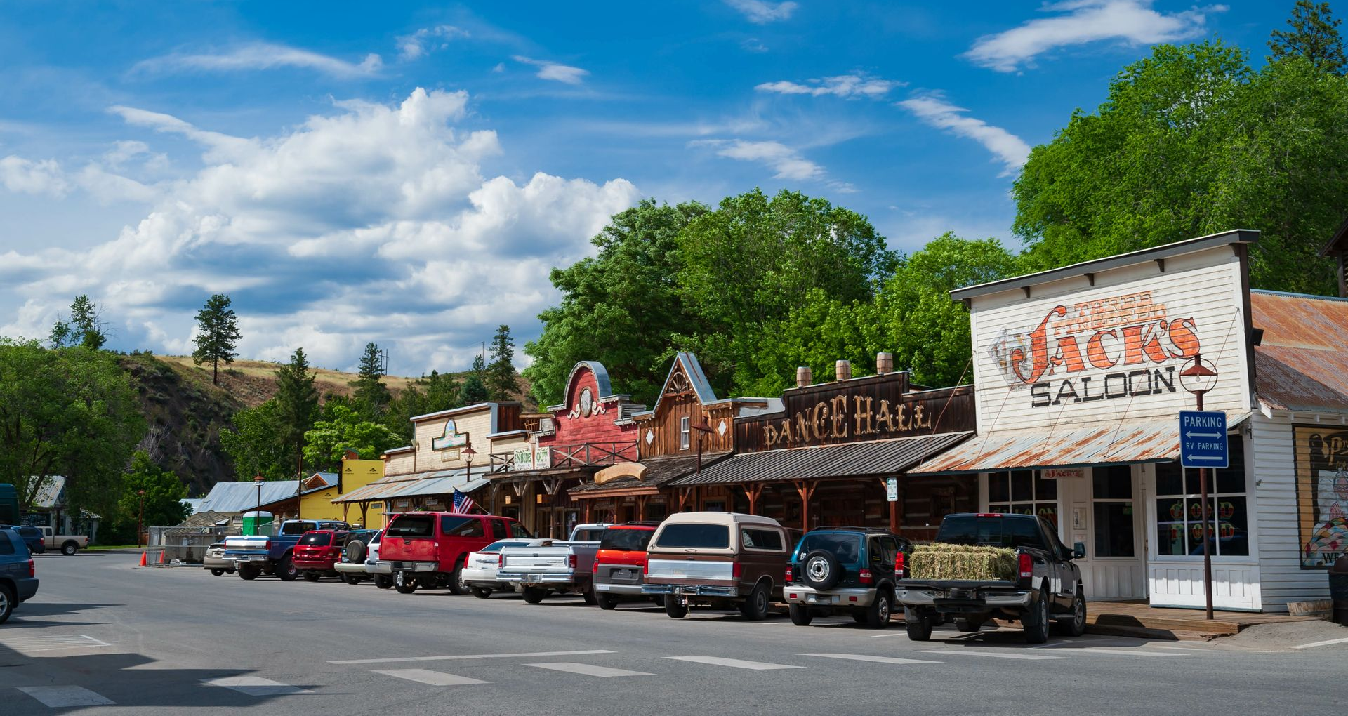Street of wooden buildings, including a saloon, with parked cars under a bright sky.