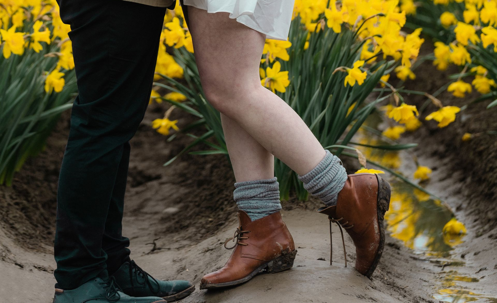 Couple standing in a field of yellow flowers. Woman wearing boots, crossing her legs. Man in black pants.