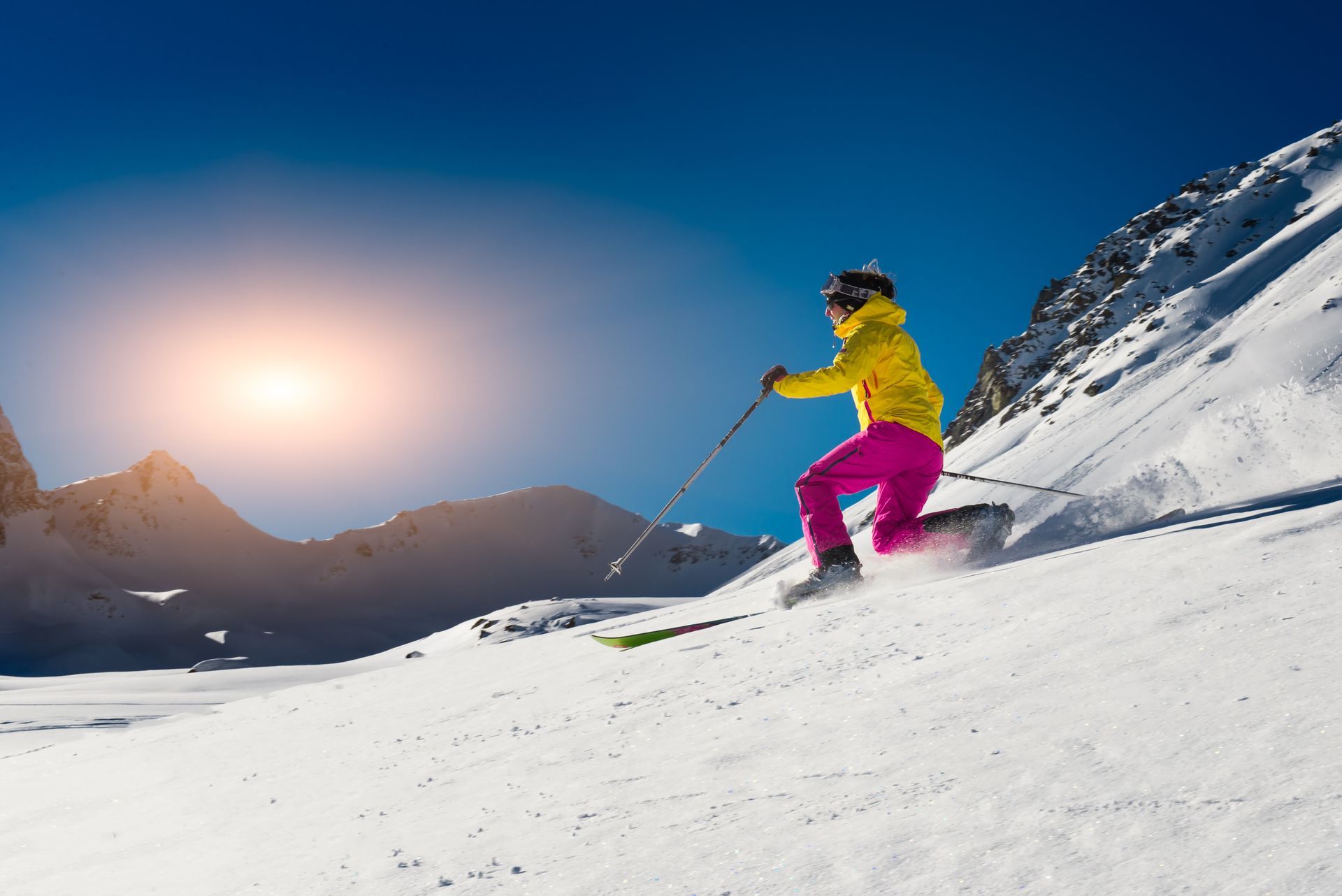 Skier in yellow jacket and pink pants carves down a snowy mountain slope under a blue sky and sun.