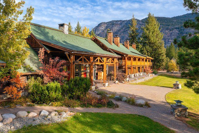 A large wooden house with a green roof is surrounded by trees and mountains.