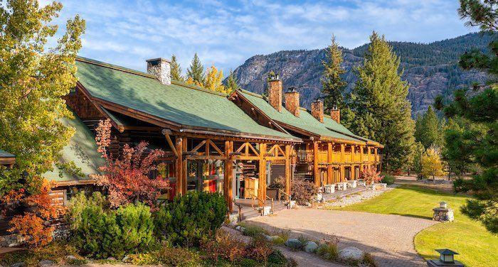 A large wooden house with a green roof is surrounded by trees and mountains.