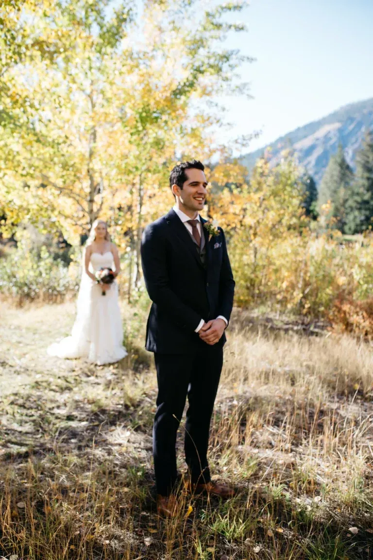 A bride and groom are standing in a field waiting for their first look.