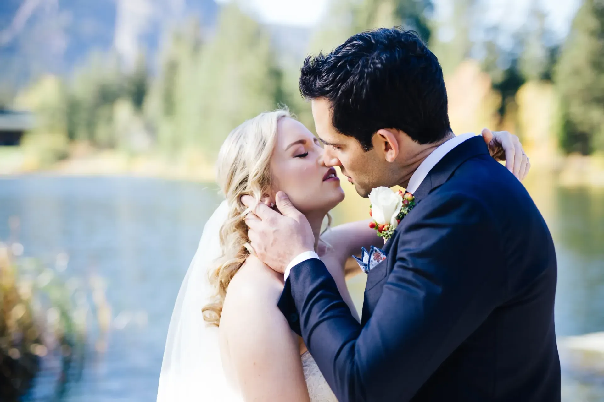 A bride and groom are kissing in front of a lake.