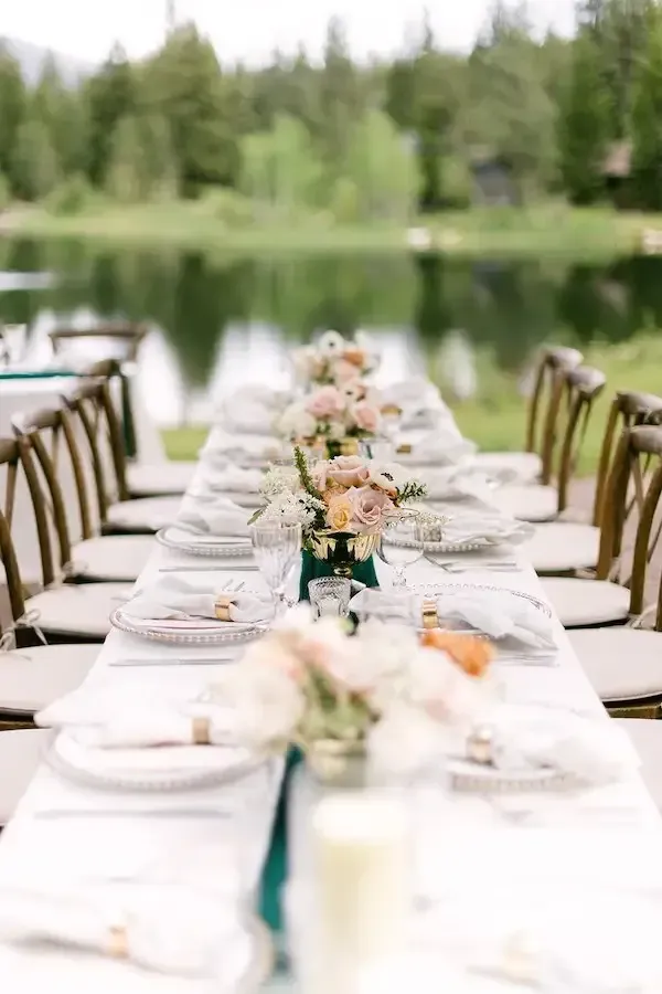 A long table with plates , utensils , and flowers in front of a lake.