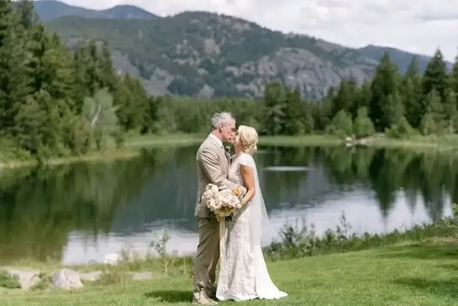 A bride and groom kissing in front of a lake with mountains in the background.