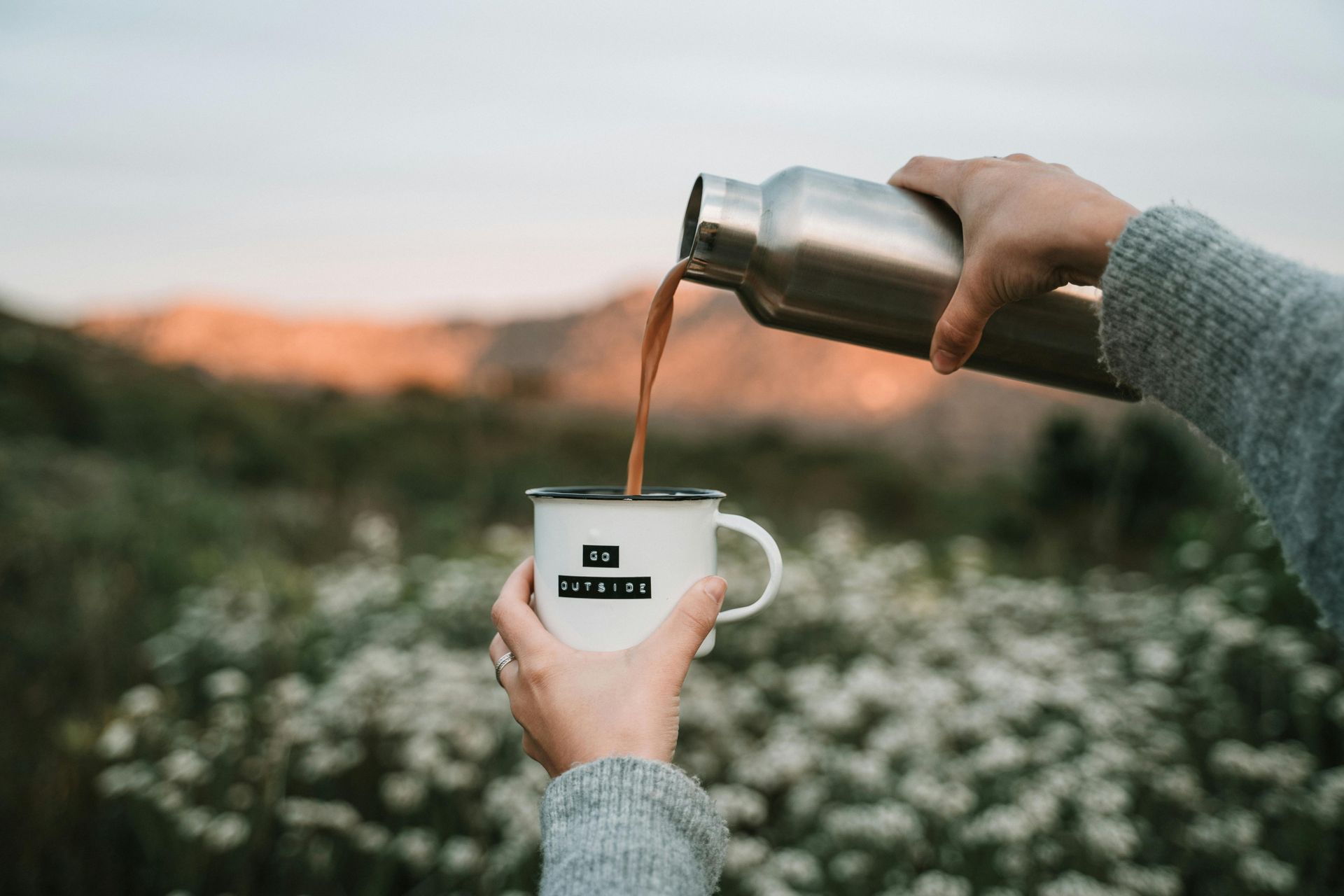 Person pouring coffee from a thermos into a mug in a field.