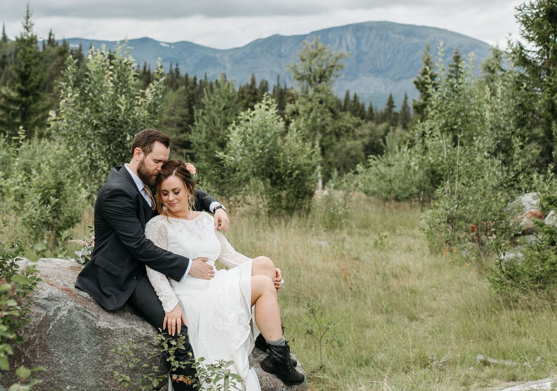 A bride and groom are sitting on a rock in a field with mountains in the background.