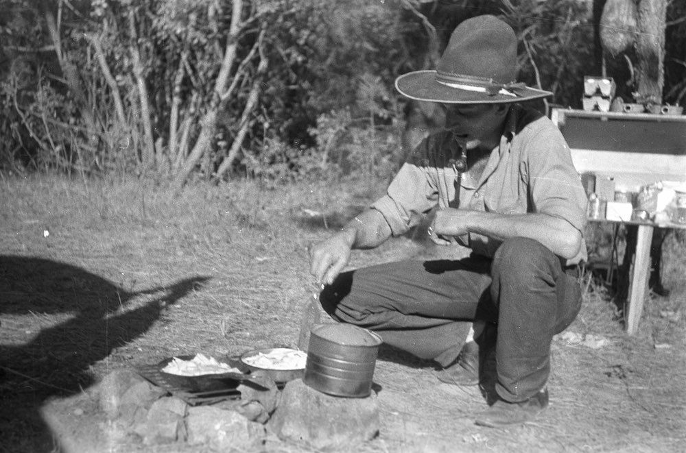 A black and white photo of a man in a cowboy hat preparing food on the ground.