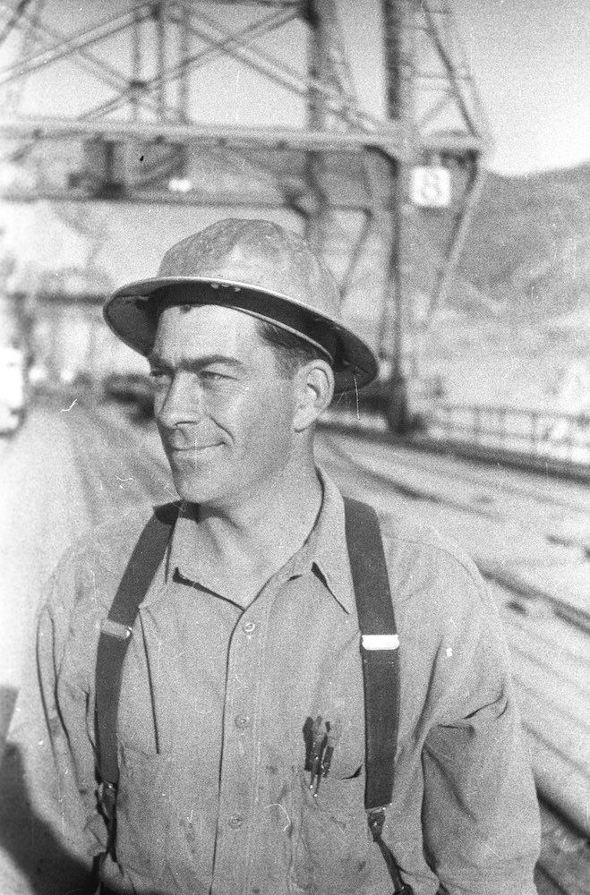 A black and white photo of a man wearing a hard hat and suspenders.