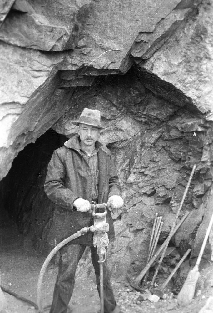 A man is standing in front of a cave holding a drill.