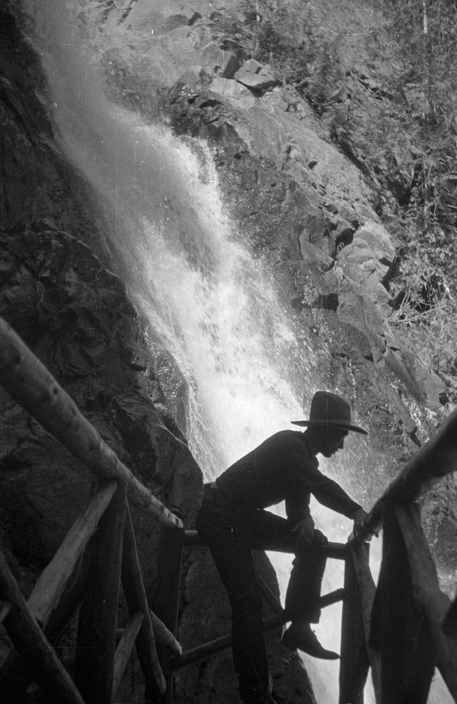 A black and white photo of a man sitting on a railing next to a waterfall.