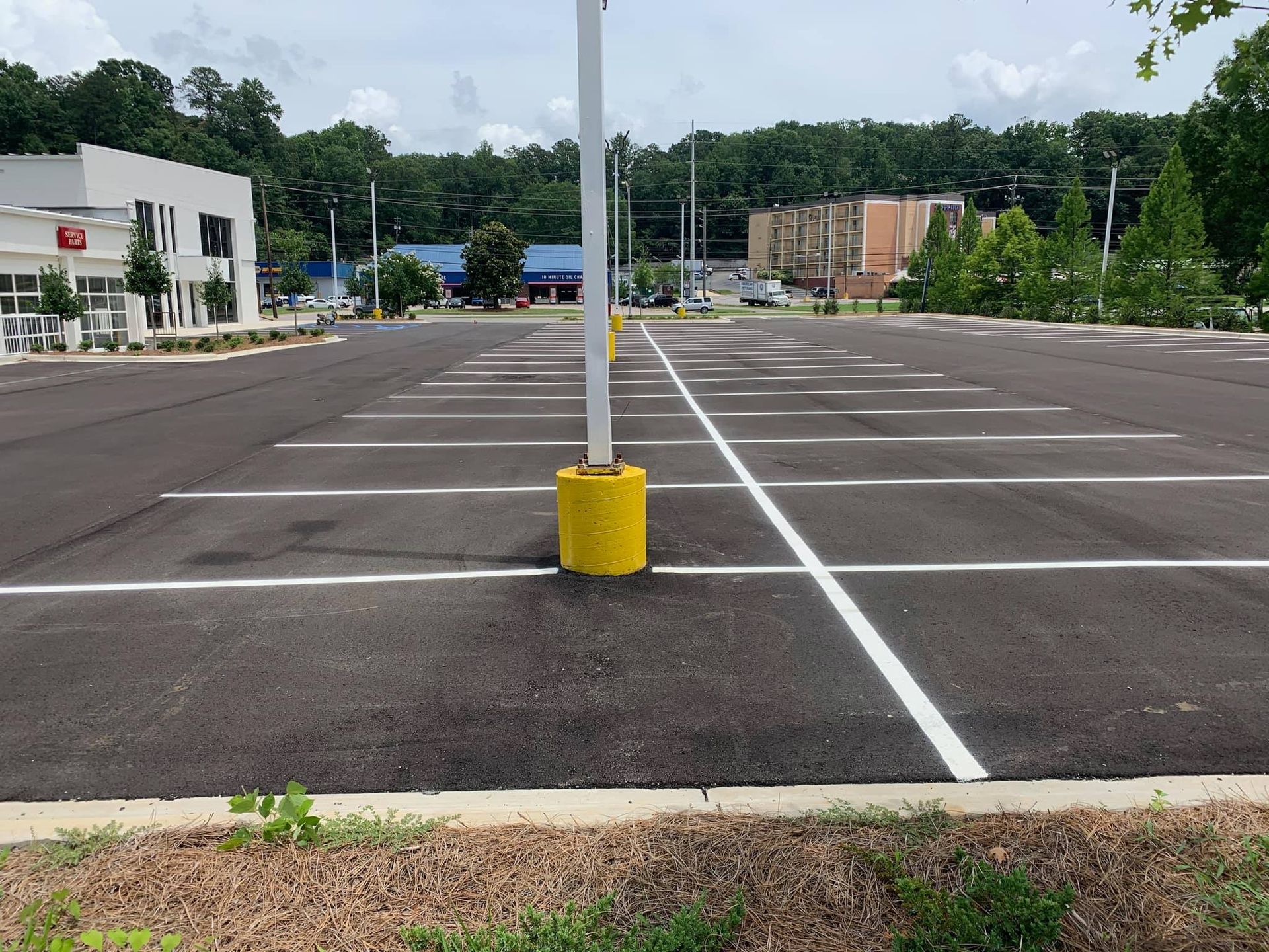 An empty parking lot with striped spaces, a yellow-based light pole, and a white building in the background.