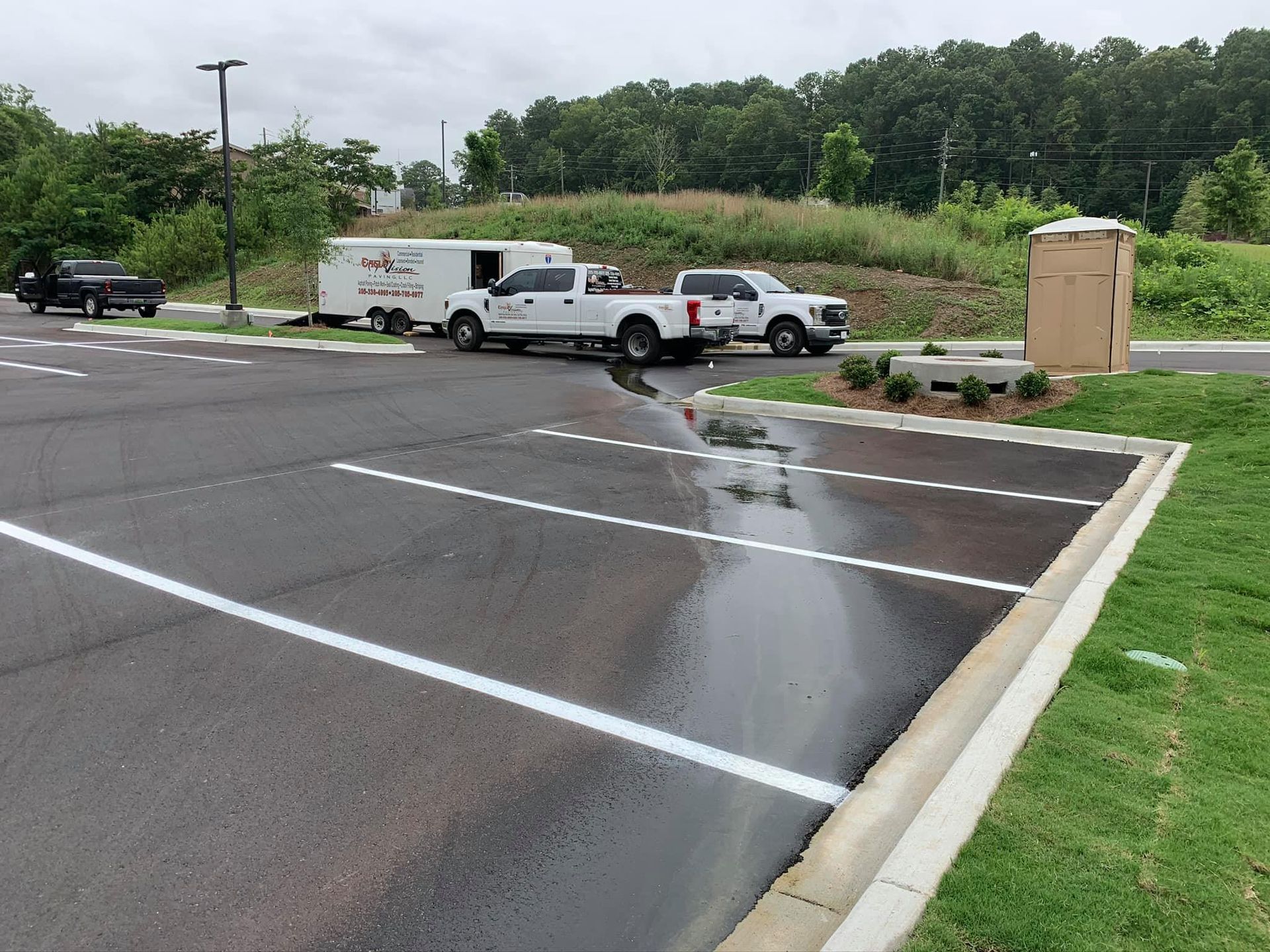 A parking lot showing two white trucks with a trailer, water pooling on the pavement, and a nearby utility enclosure.