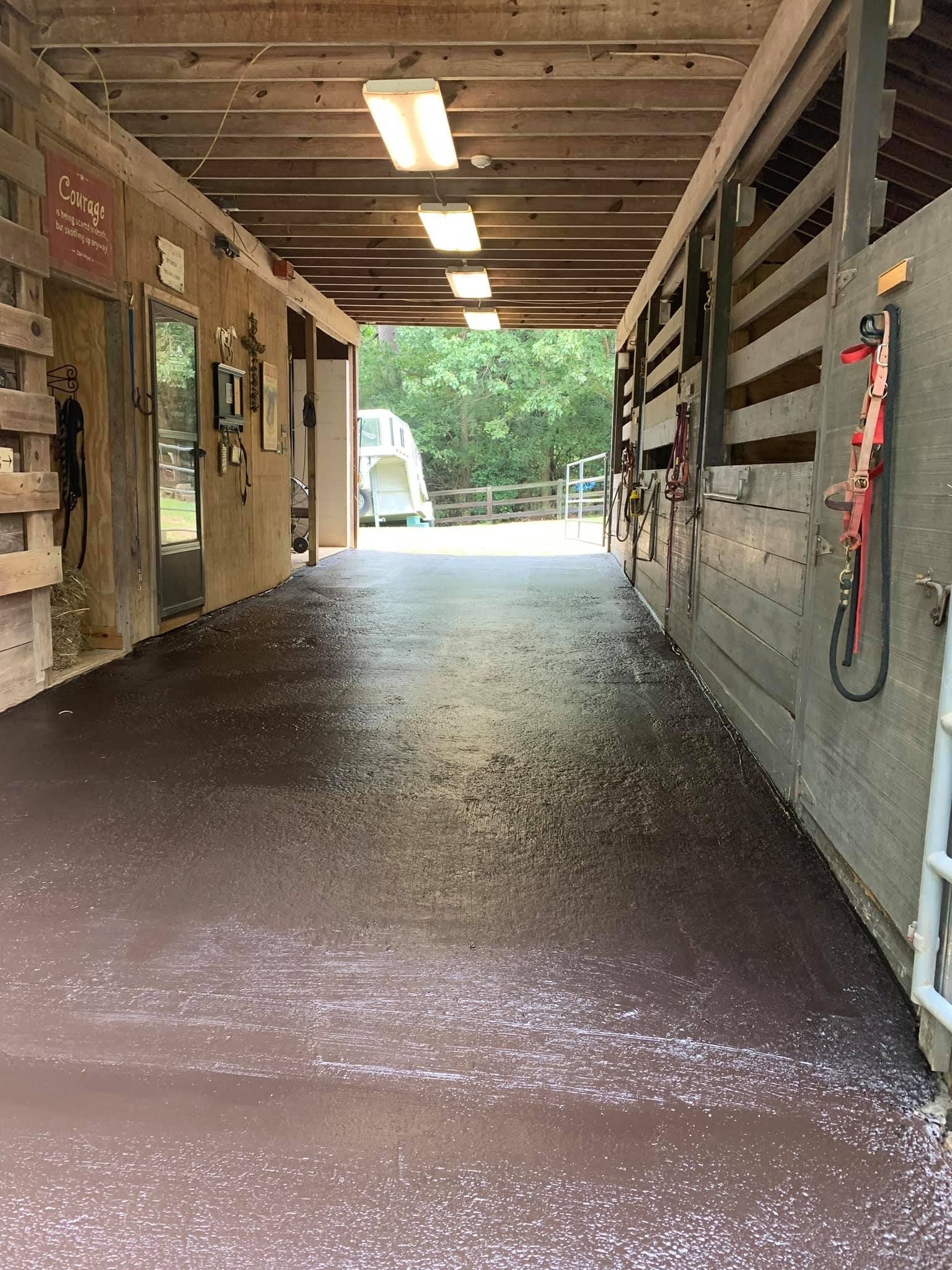 A covered outdoor aisle between horse stalls with a dark, speckled concrete floor and white ceiling lights.