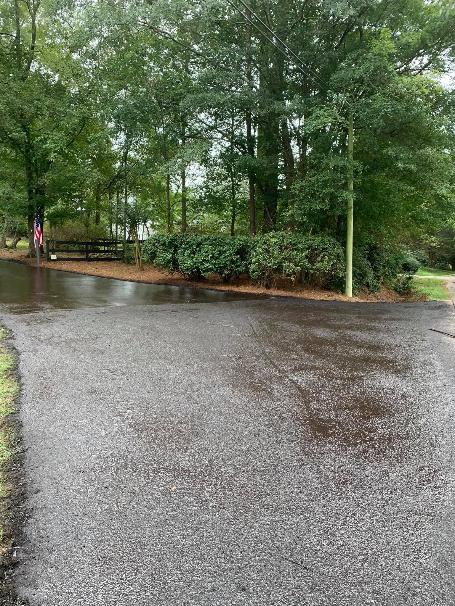 A wet, gravel driveway transitions to a paved surface, leading toward a line of lush green trees and a small wooden fence.