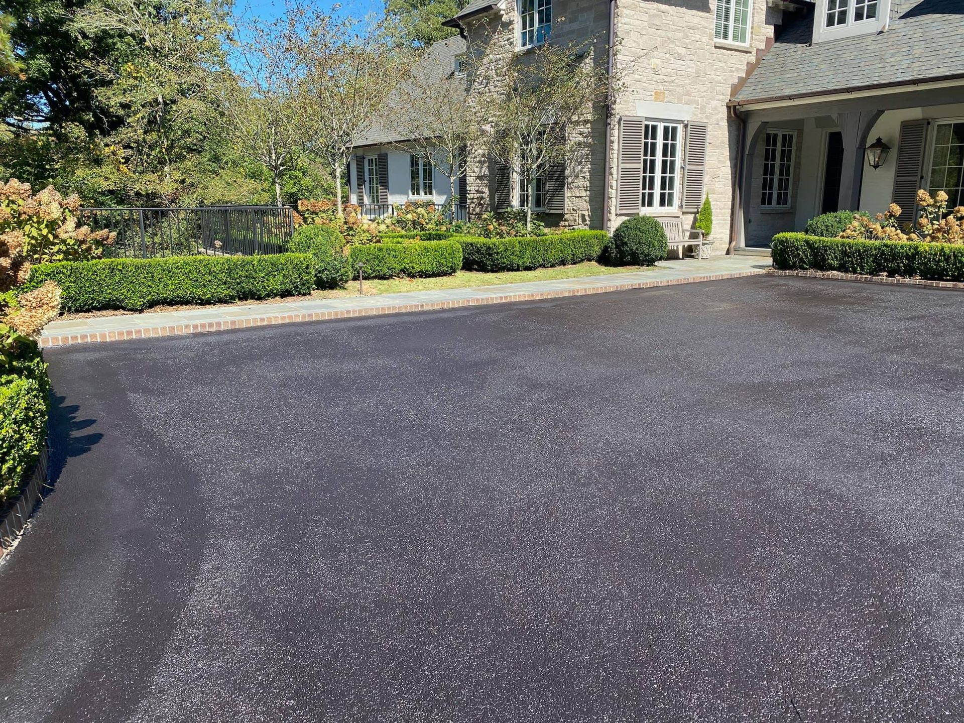 A black asphalt driveway in the foreground leading to a stone house with manicured green hedges.