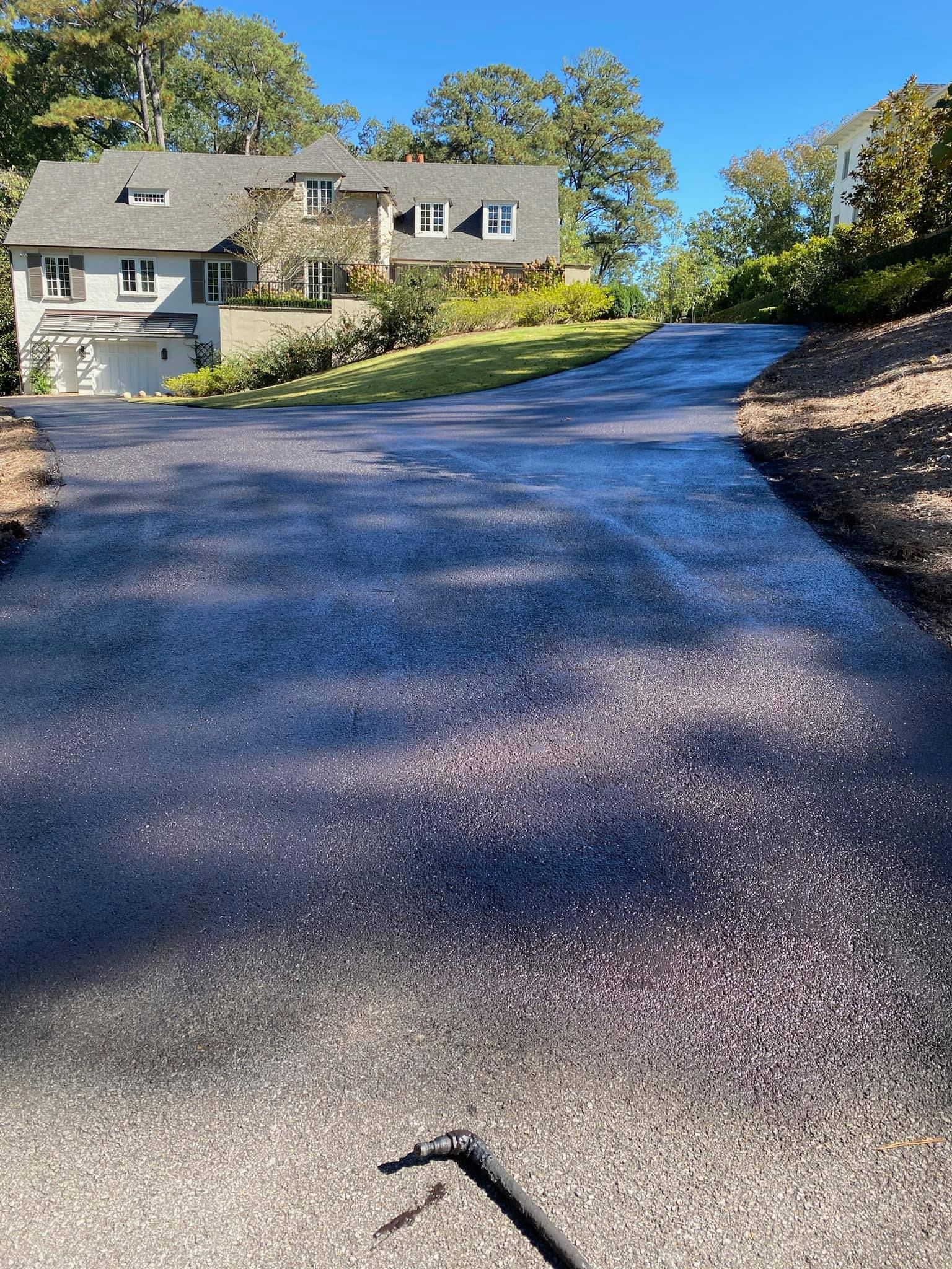 A paved driveway, freshly coated with a dark sealant, leading toward a stone-accented house on a sunny day.