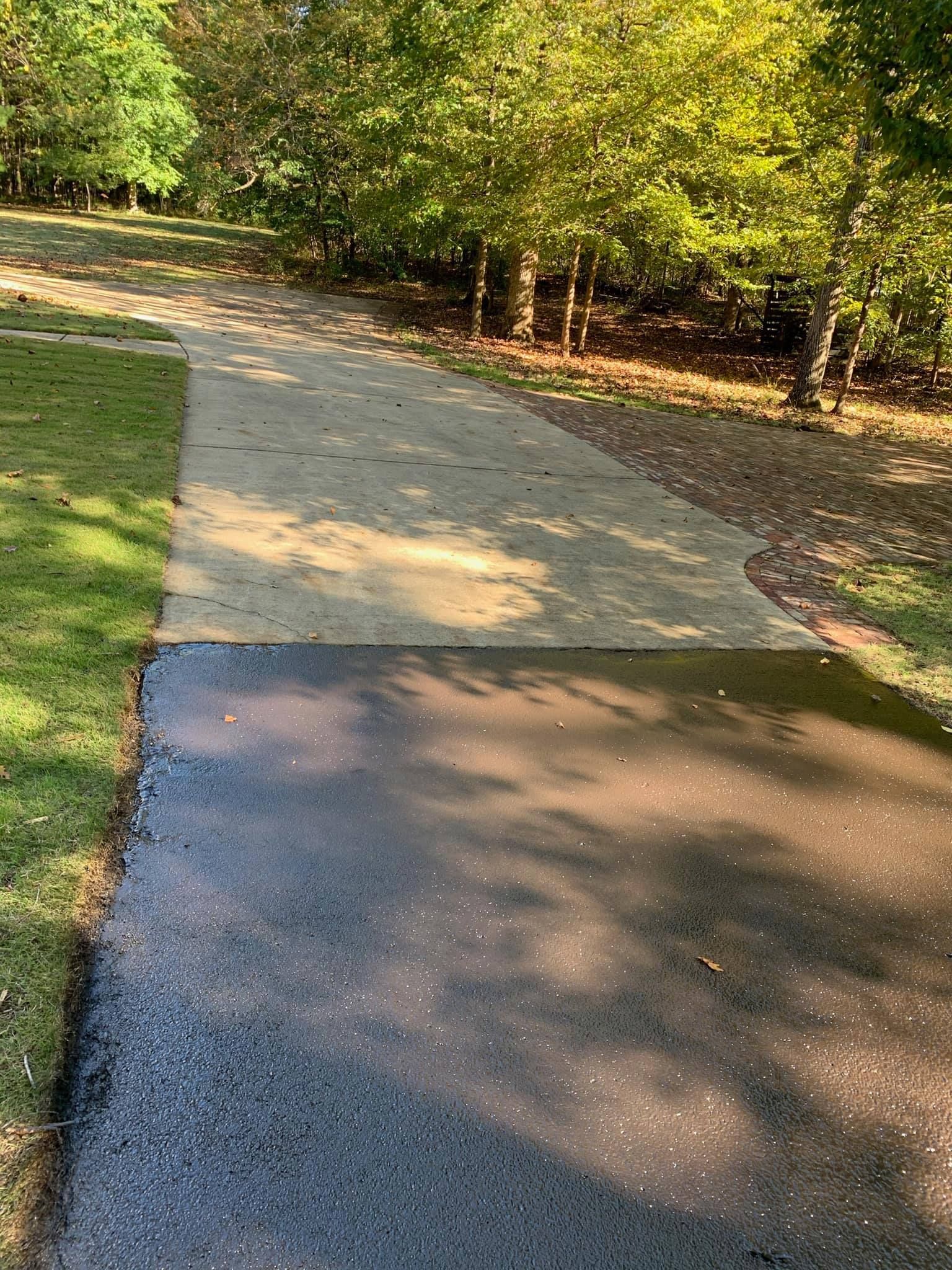 A concrete driveway transitions from a dry, light-colored section to a wet, dark section near a treeline.