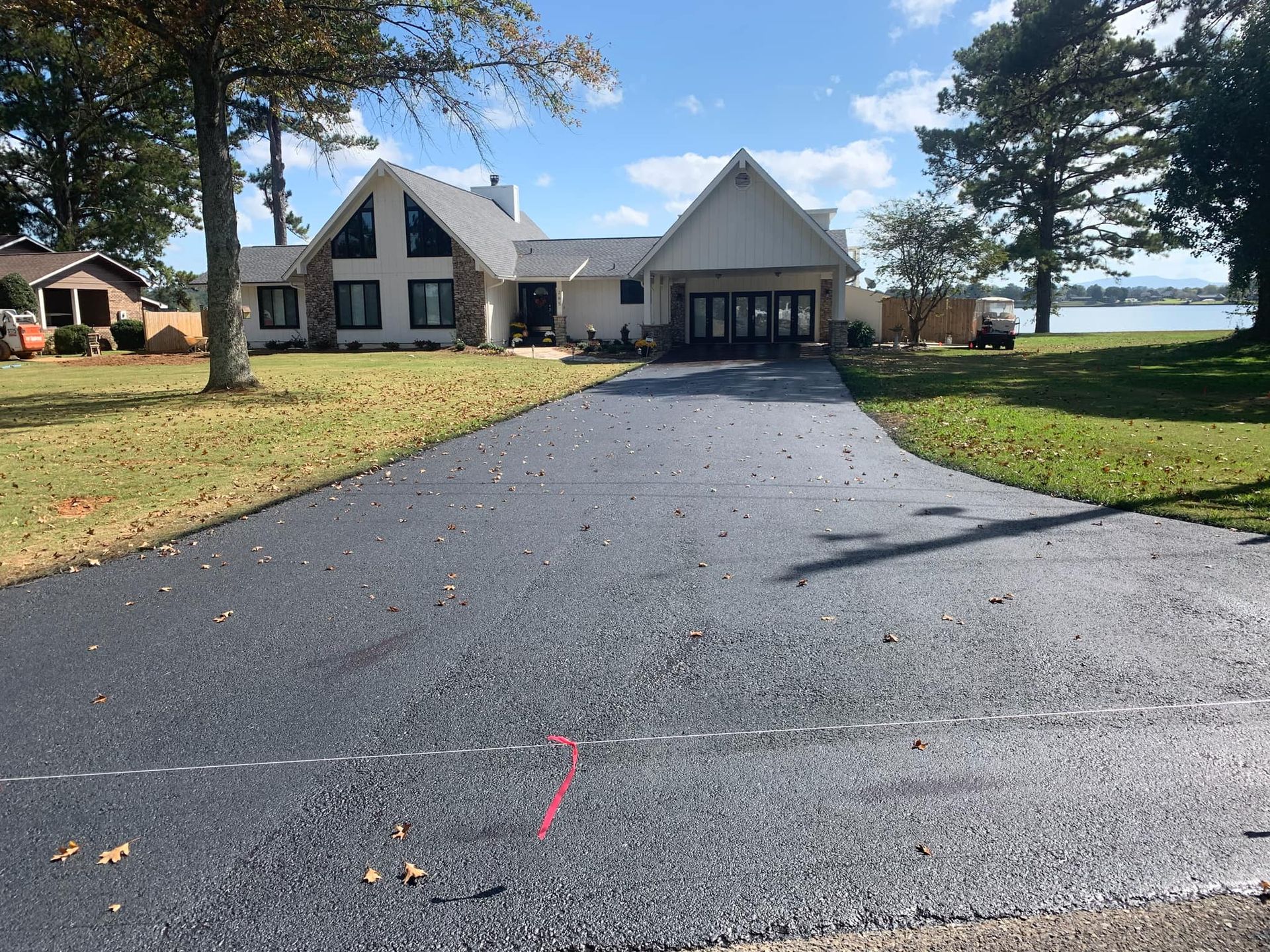 A freshly paved black asphalt driveway leads toward a two-story light-colored house with a yard on a sunny day.