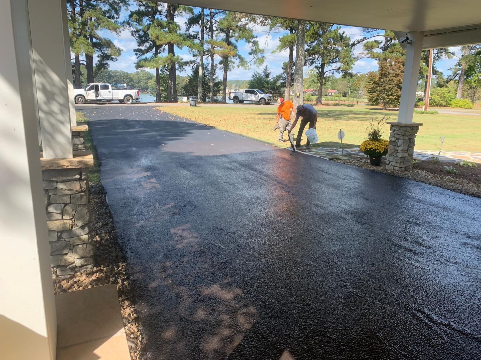 Two people work on a freshly paved, dark asphalt driveway outside a home on a sunny day.