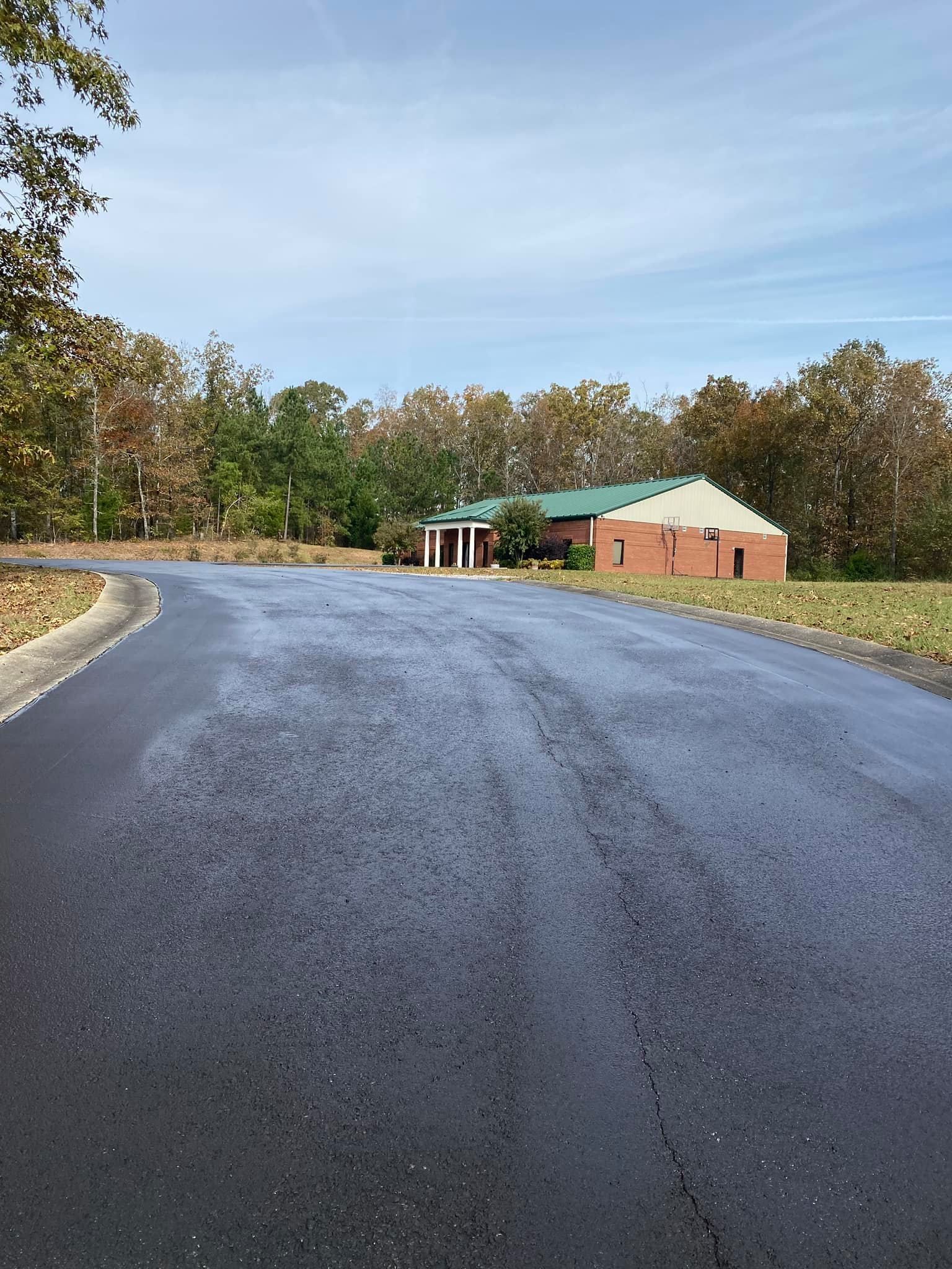 A newly paved black asphalt road leads toward a brick building with a green roof, surrounded by trees on a sunny day.