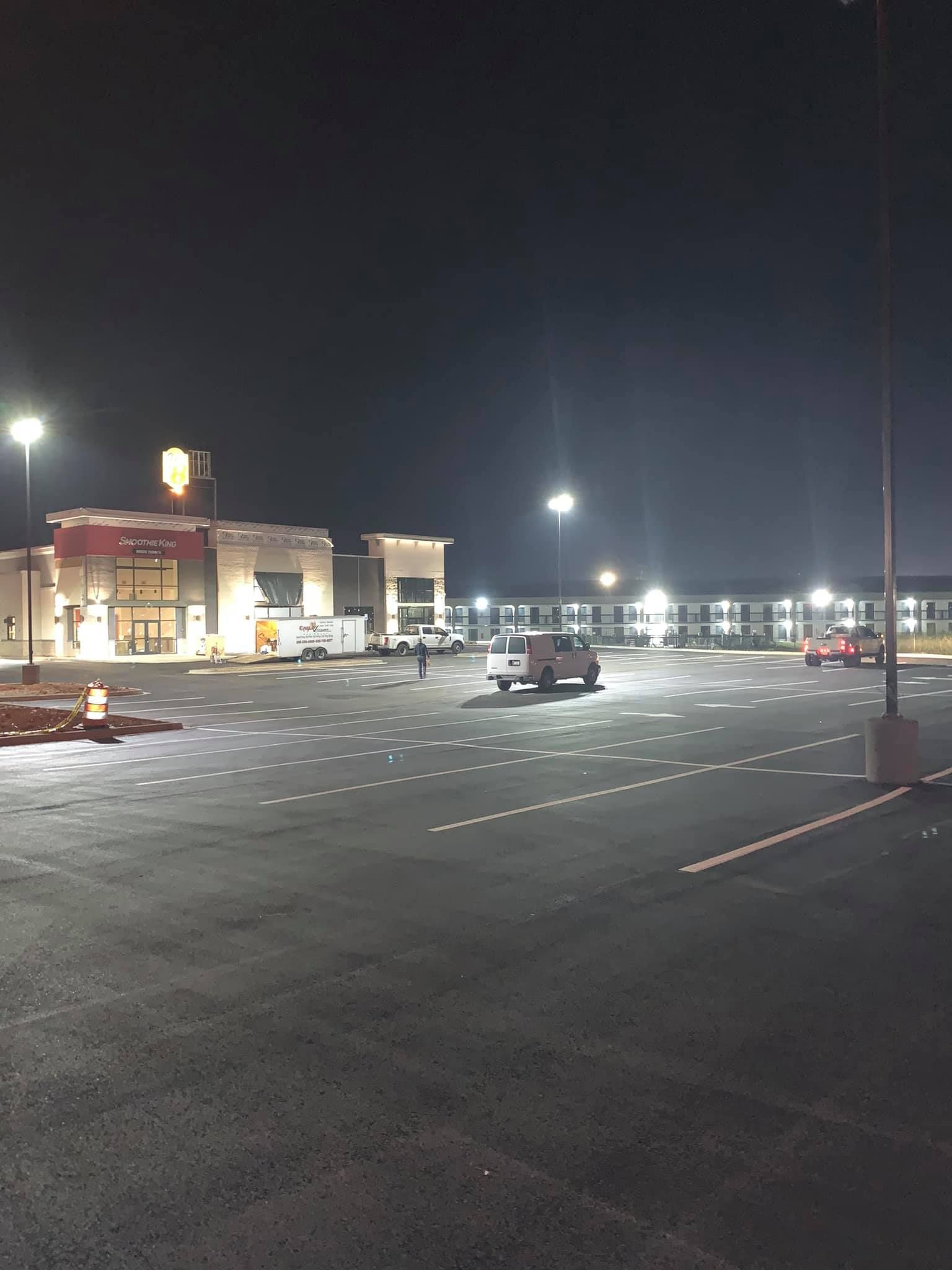 A night view of a brightly lit parking lot in front of a hotel building with a few parked vehicles.
