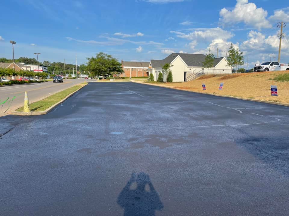 A freshly paved asphalt parking lot sits next to a grassy embankment and a suburban building under a blue sky.