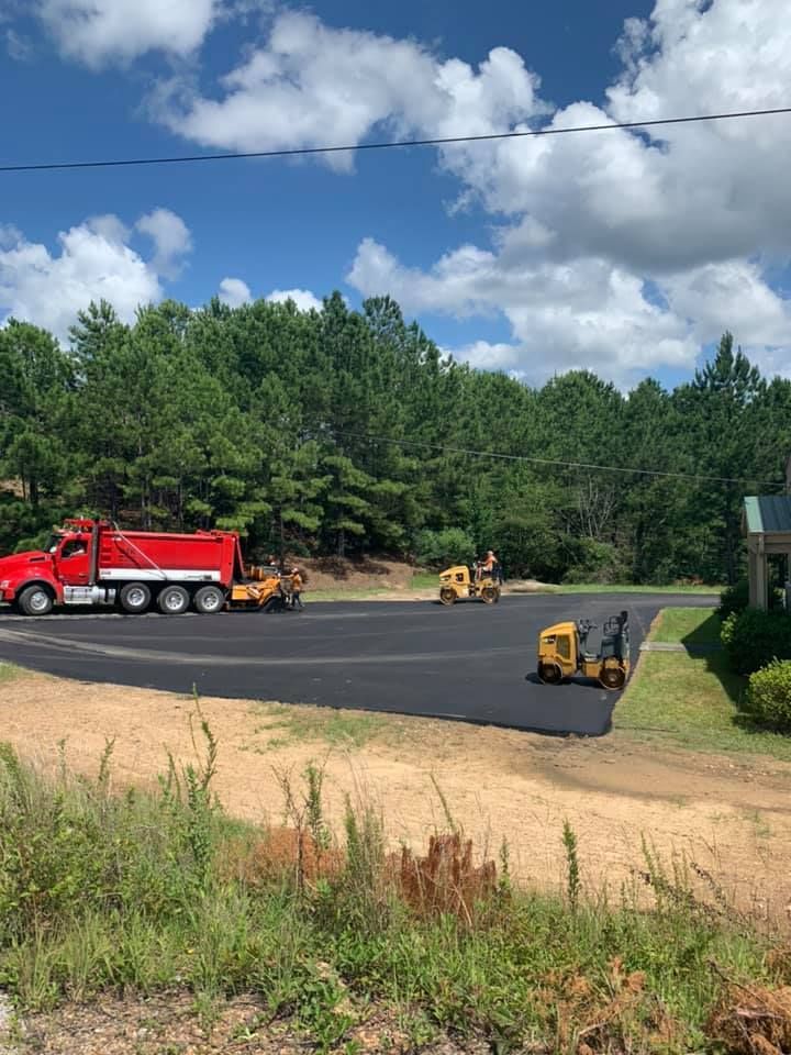 A red dump truck and construction equipment paving a new asphalt lot in a wooded, sunny area.