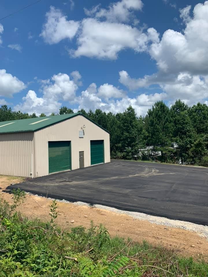 A tan metal storage building with a green roof and two green roll-up doors, set beside a freshly paved asphalt driveway.