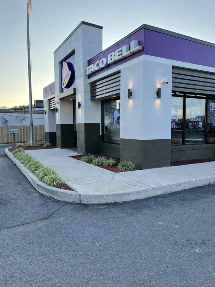 A modern Taco Bell restaurant with white and dark gray walls, purple accents, and a sidewalk entrance at sunset.