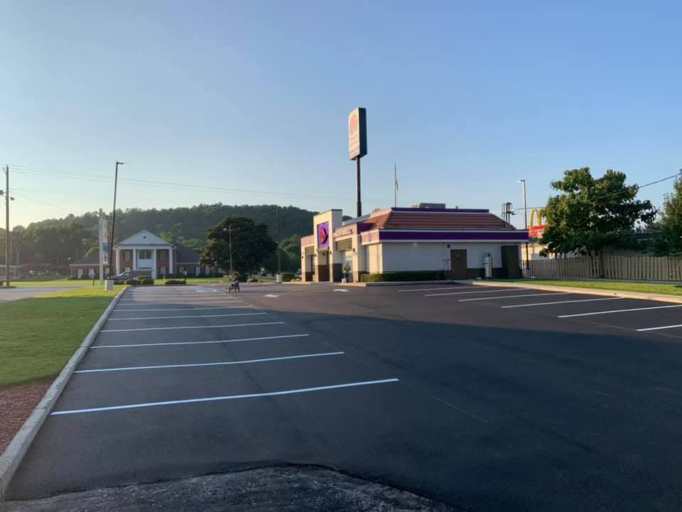 A Taco Bell restaurant with a parking lot in the foreground, set against a backdrop of trees and a clear blue sky.