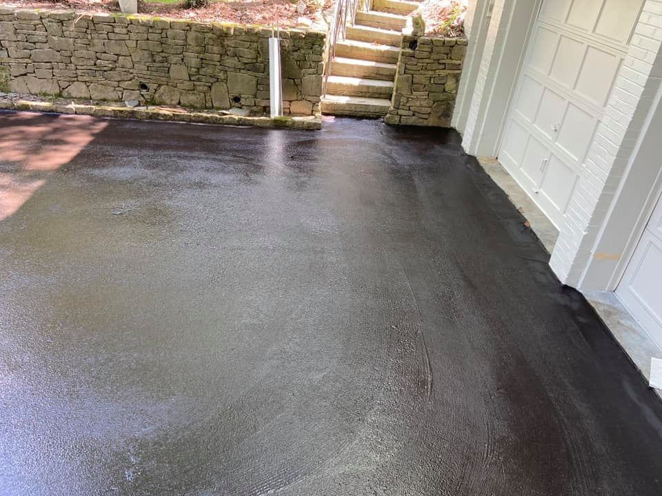 A freshly sealed black asphalt driveway next to a white garage, with a stone retaining wall and steps in the background.