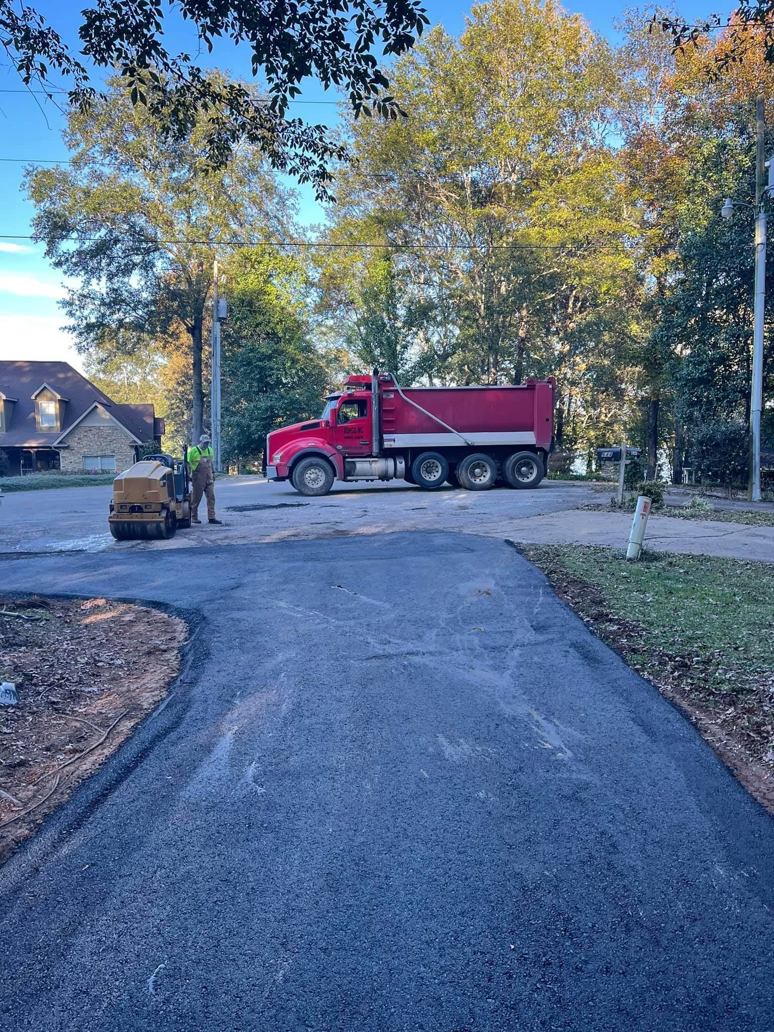 A red dump truck and a small yellow roller on a residential street during an asphalt paving project.