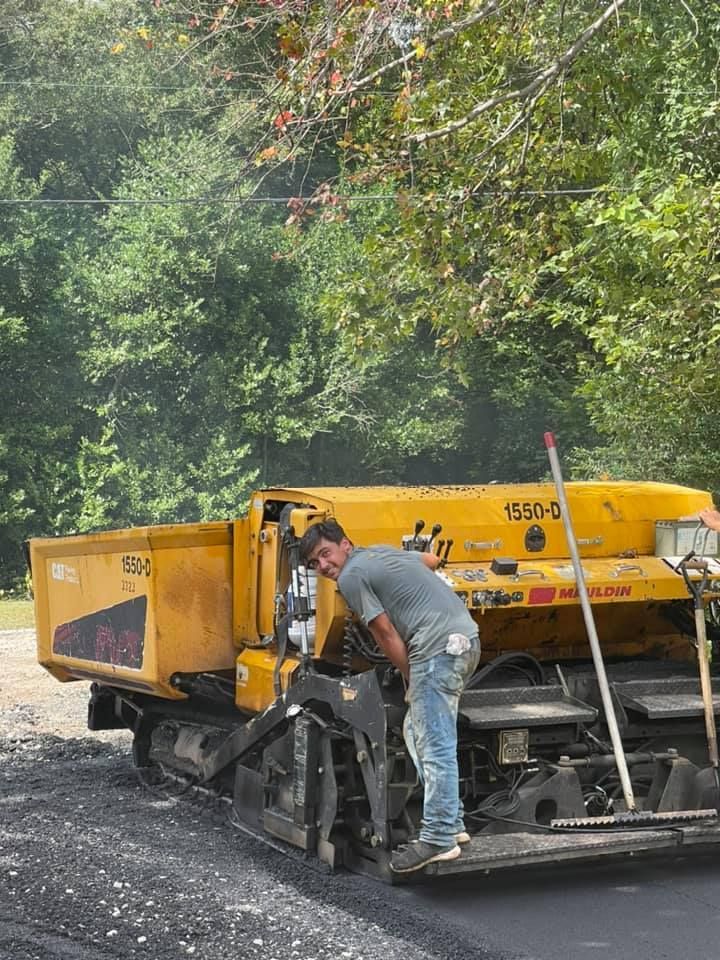 A person in work clothes stands on a yellow construction paver on a paved road surrounded by trees.
